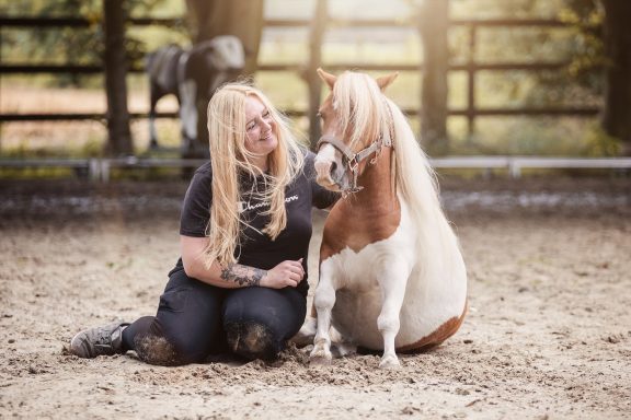 Frau mit langen blonden Haaren sitzt neben einem kleinen Pferd in einer Reitarena.