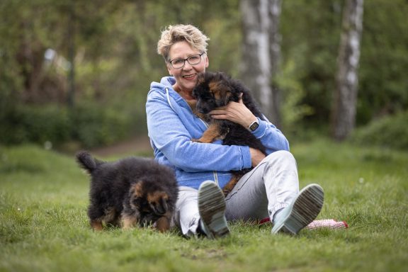 Eine lächelnde Frau sitzt im Gras und hält zwei Welpen, einer schnüffelt am Boden.